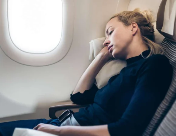 Frau schläft entspannt auf einem Fensterplatz im Flugzeug mit angelehntem Kopf und Sicherheitsgurt angeschnallt. Gehörschutz beim Fliegen kann helfen, störenden Lärm auszublenden und Ohrendruck zu vermeiden.