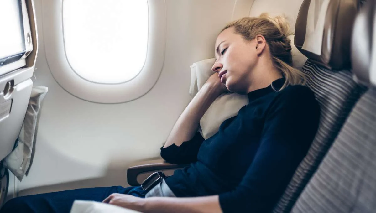 Frau schläft entspannt auf einem Fensterplatz im Flugzeug mit angelehntem Kopf und Sicherheitsgurt angeschnallt. Gehörschutz beim Fliegen kann helfen, störenden Lärm auszublenden und Ohrendruck zu vermeiden.