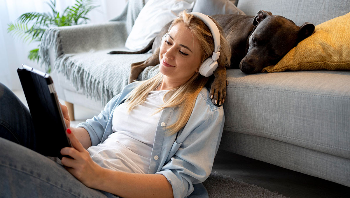 Frau mit Kopfhörern und Tablet, neben ihr liegt ein Hund auf dem Sofa - Online-Hörtest für die schnelle Überprüfung des Gehörs in wenigen Minuten, ideal für einen ersten Überblick über die Hörfähigkeit.